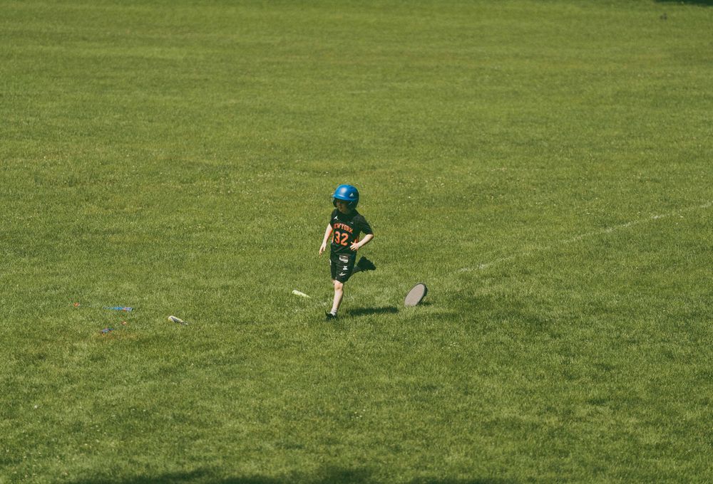 Young child in green uniform and helmet running on a grass field during a youth football game.