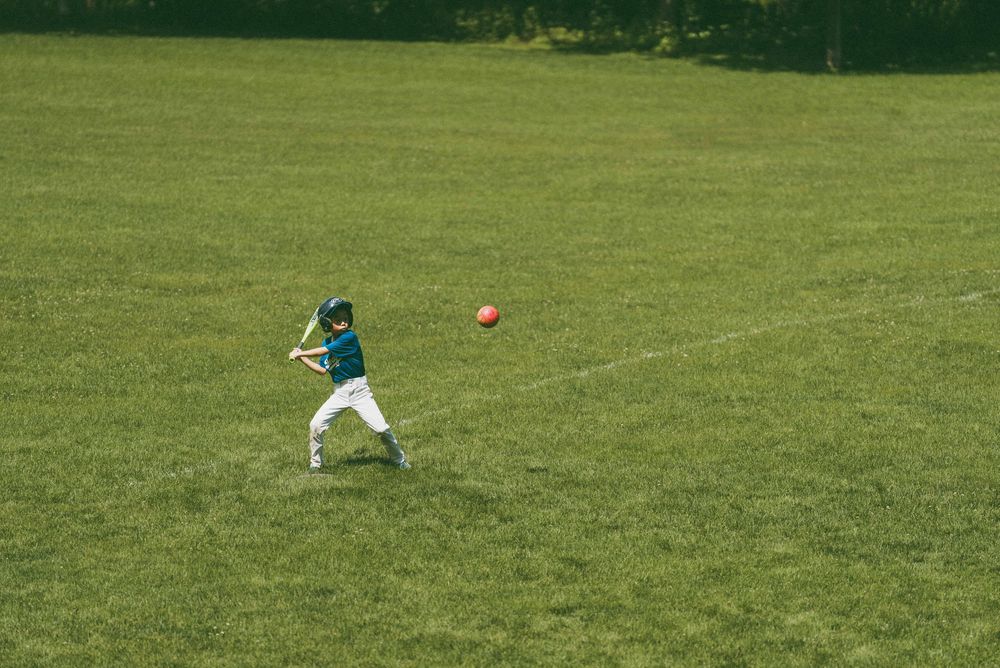 Young baseball player in teal uniform swinging bat at red ball on sunny green field.
