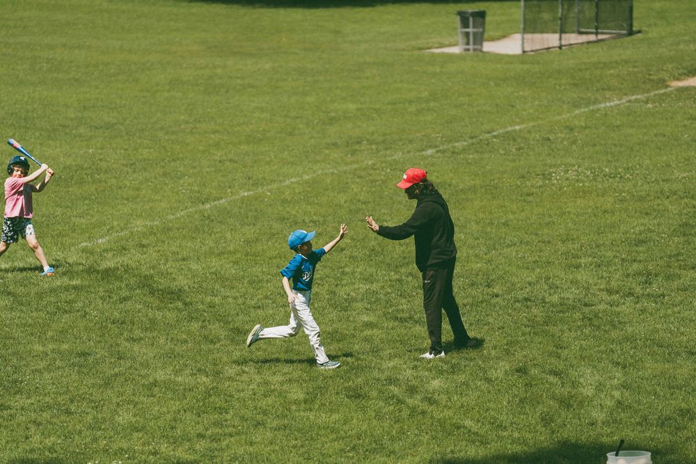Children playing baseball on a green field, with a coach in a red cap giving a high five to a young player.