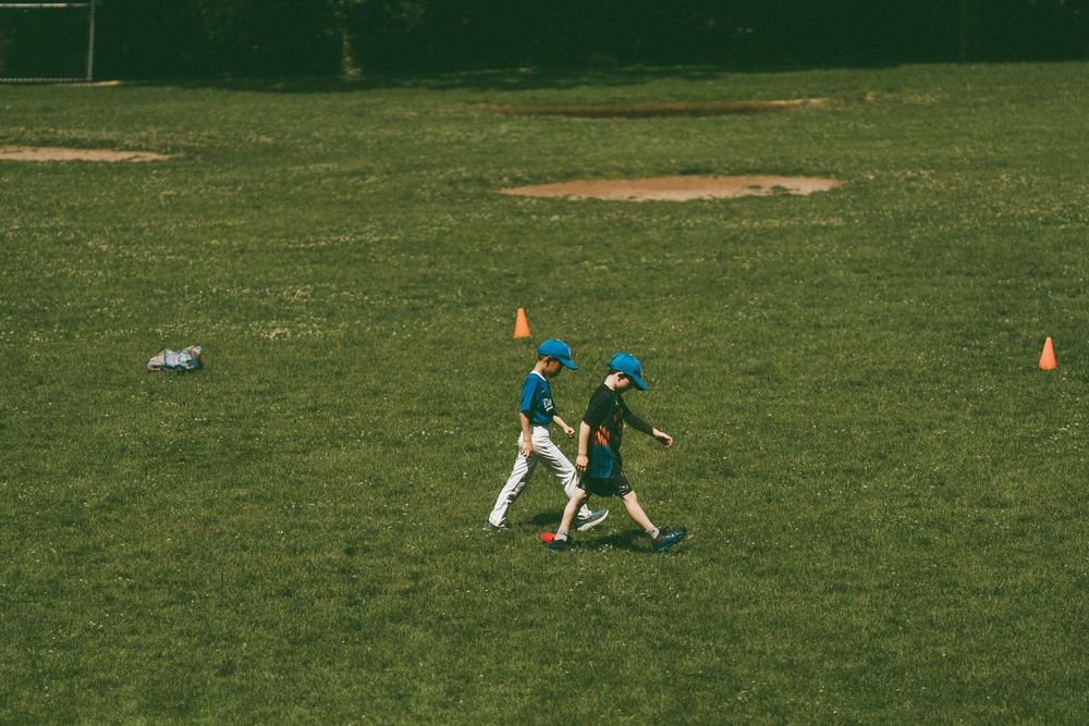 Two young baseball players in blue helmets walk across a sunny green field during practice drills.