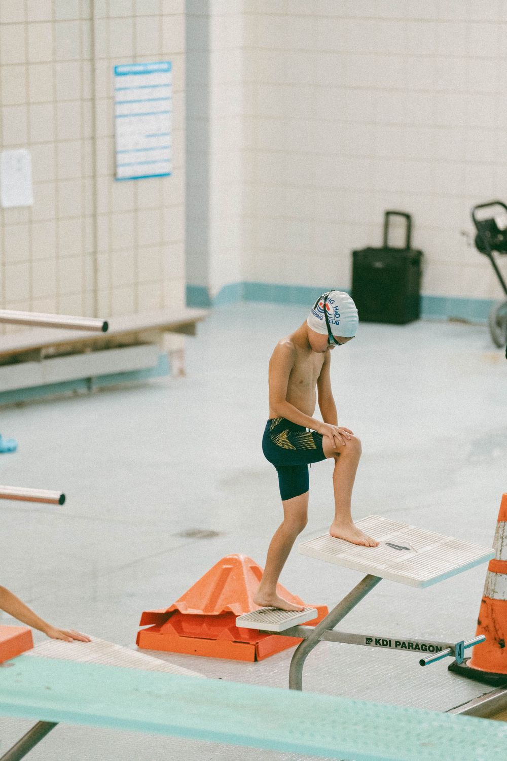 Young competitive swimmer in cap and goggles stands on starting block at indoor pool, ready to race.