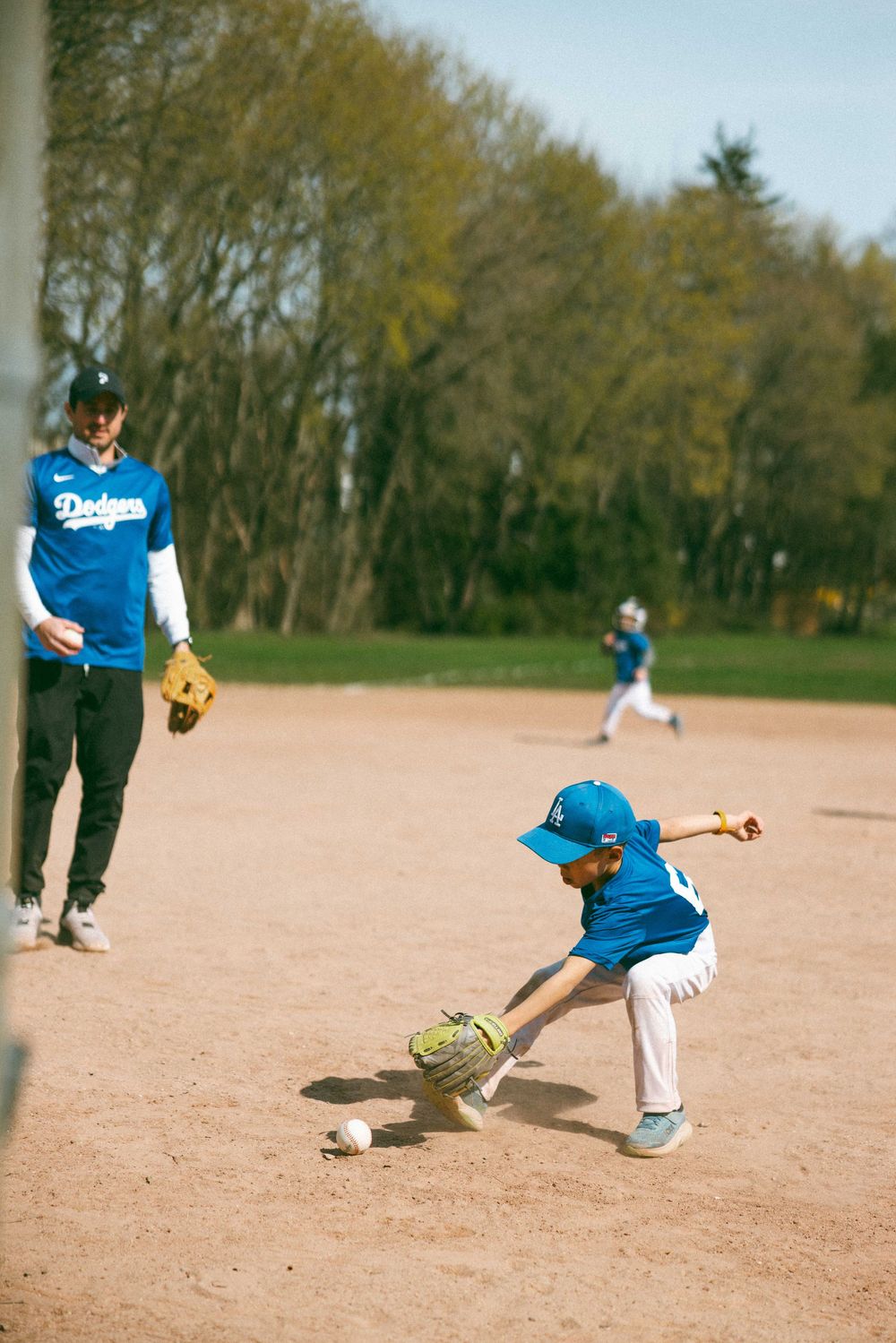 Young Dodgers player fielding a ground ball during youth baseball practice with coach watching nearby.