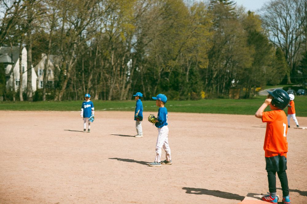 Young baseball players in blue uniforms practice fielding drills on a sunny dirt baseball diamond surrounded by green trees.