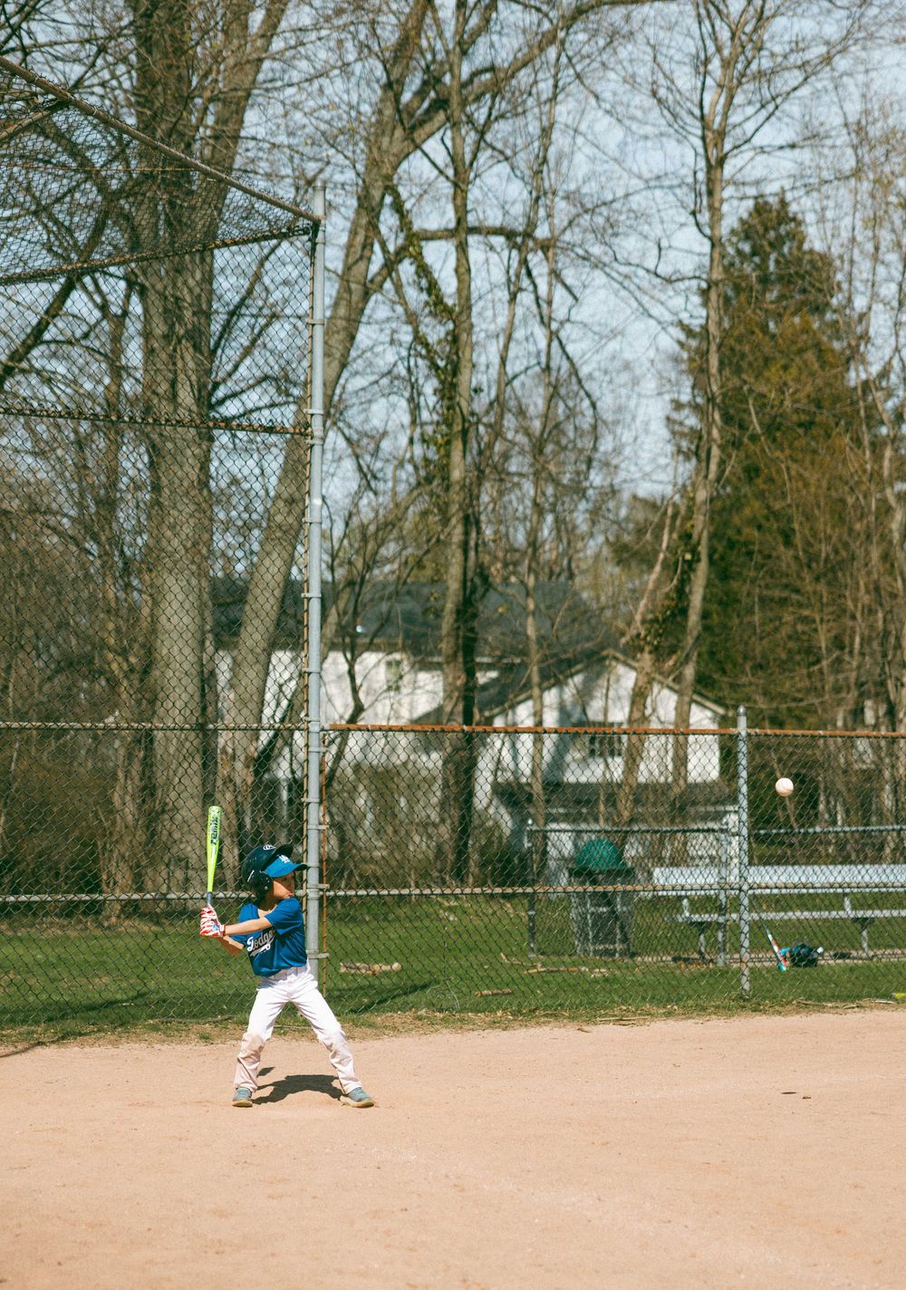 Young baseball player in blue jersey swinging bat on dirt field with chain-link fence and bare trees in background.