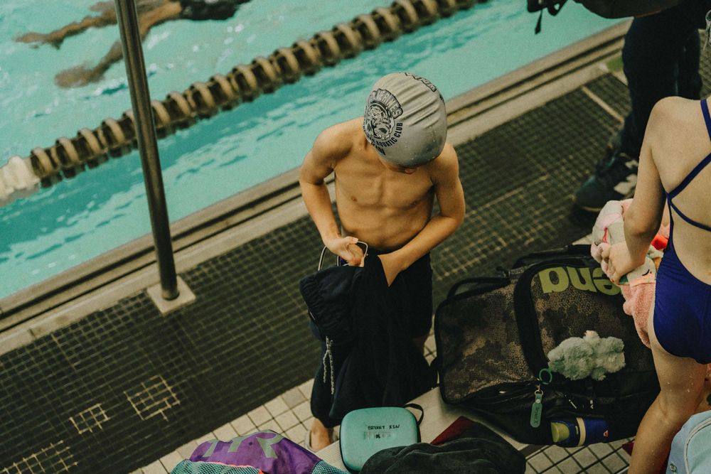 A person wearing a swim cap sits poolside with a bag and phone during a swimming session.