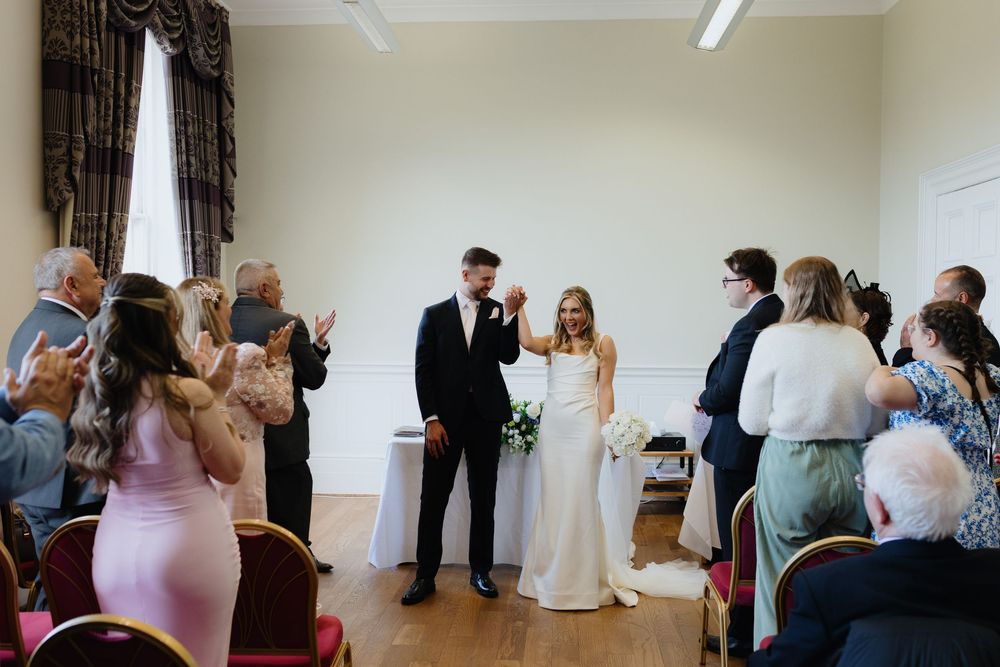 Wedding ceremony guests stand in a bright indoor venue with white walls and wooden floors.