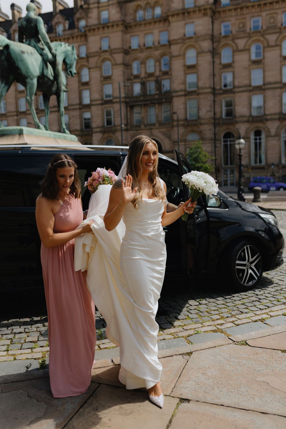 Wedding party members in pink and white dresses walk together on a cobblestone street outside a historic building.