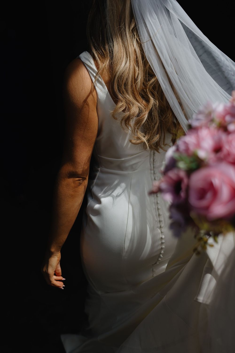 Close-up of bride in elegant white wedding dress with flowing veil and pink rose bouquet in moody lighting.