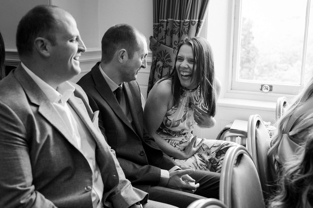 A candid black and white photograph shows people laughing together while seated on a couch by a window.