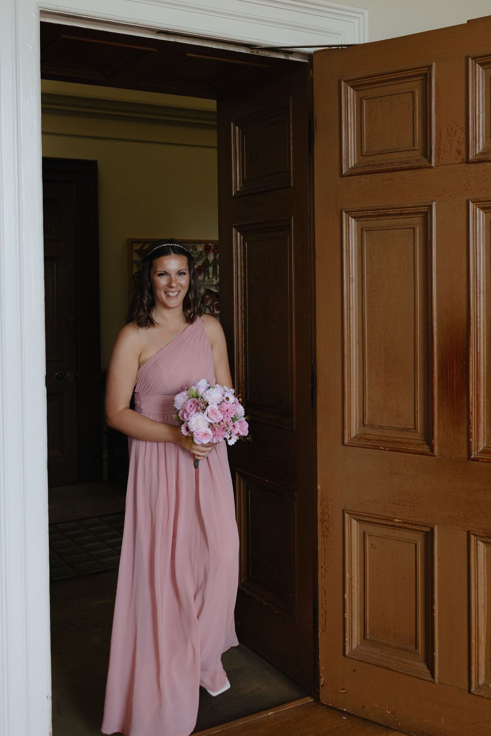Bridesmaids in pink dresses stand in a wood-paneled hallway holding bouquets.