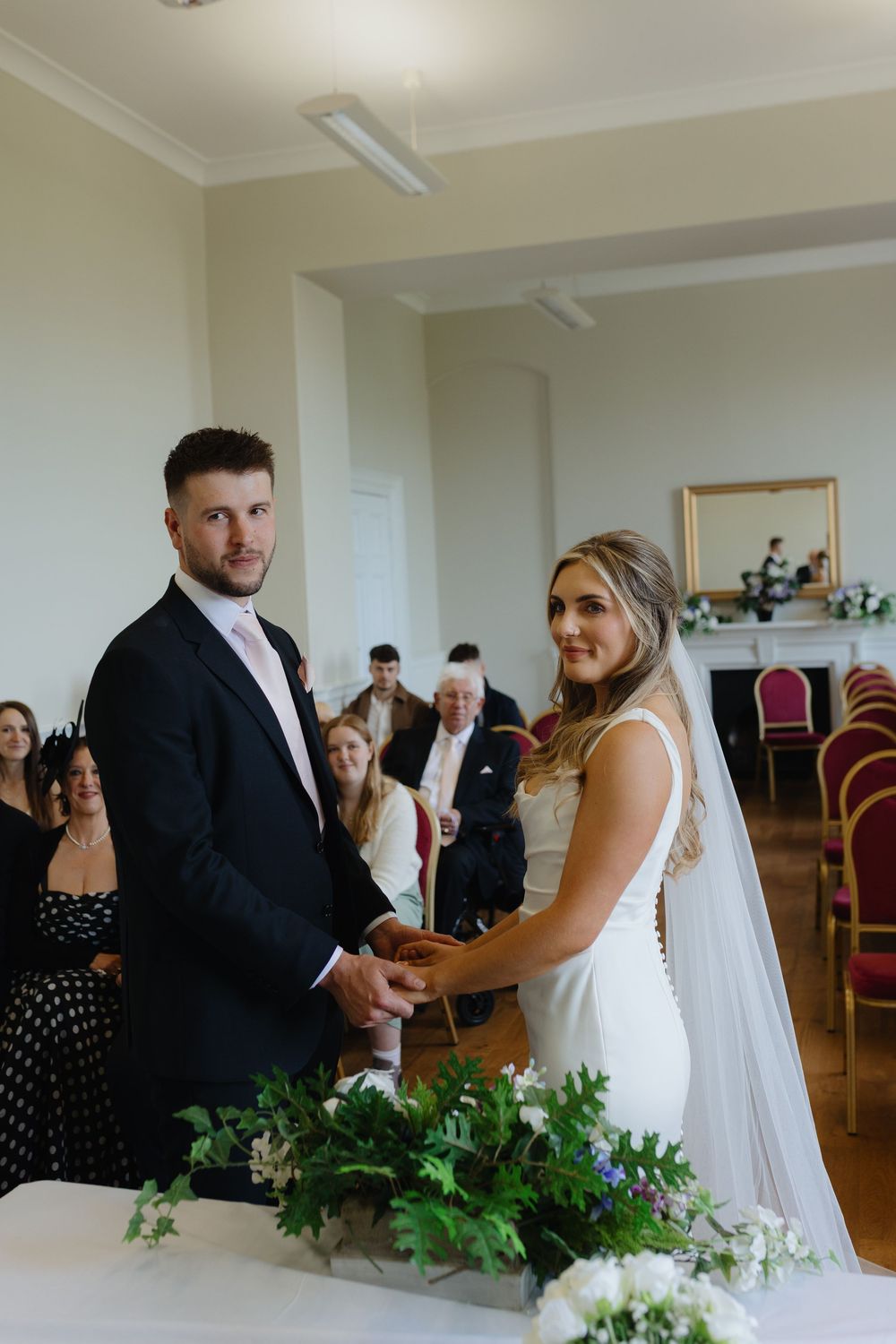 Wedding ceremony in a bright indoor venue with white floral decorations and guests seated in rows.