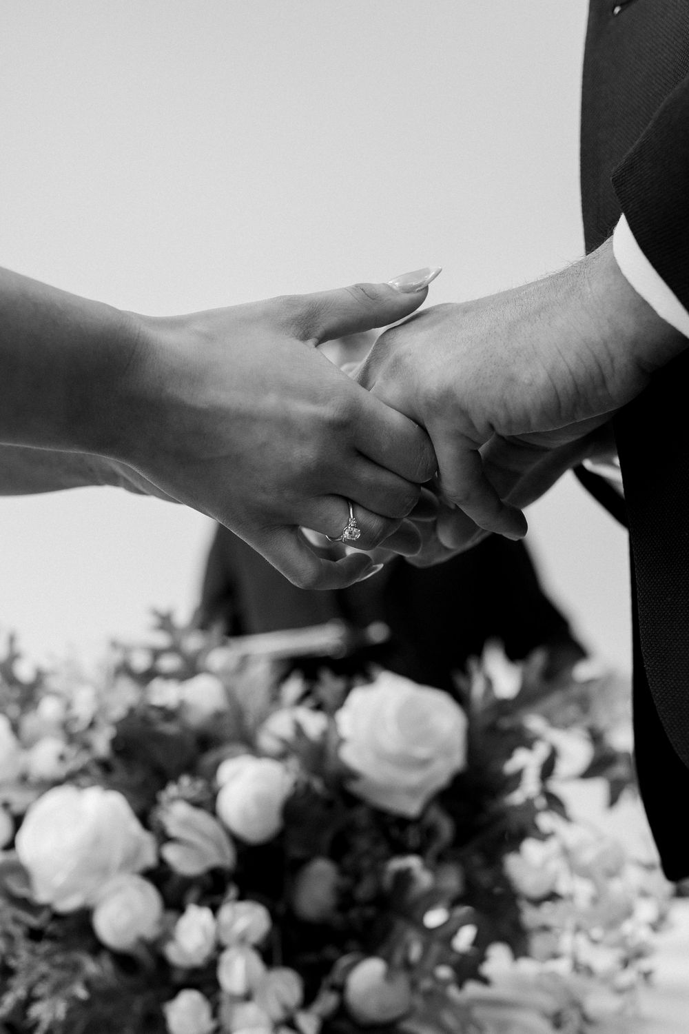 Two hands exchanging rings during a wedding ceremony with blurred white flowers in the background.