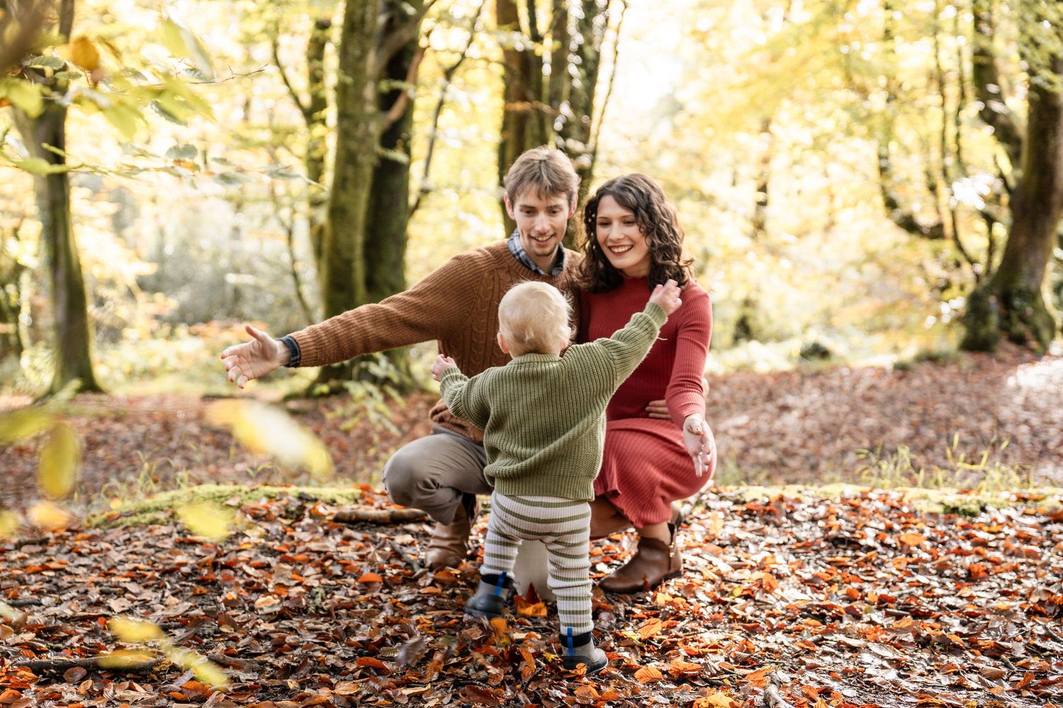 A small child plays in fallen autumn leaves while wearing a brown sweater in a wooded area.