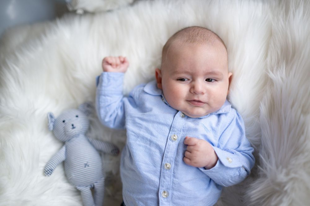 A baby in a light blue outfit lies on a white fluffy blanket with small plush animal toys.