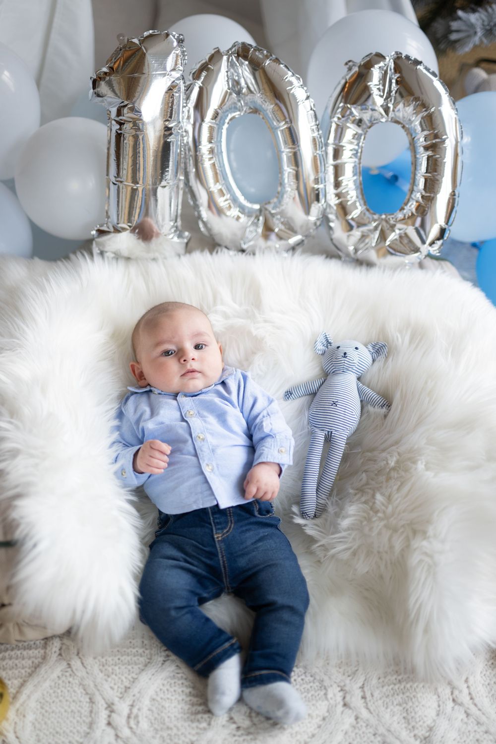 Baby lying on white fluffy blanket with silver balloon decorations and stuffed elephant toy in nursery setting.