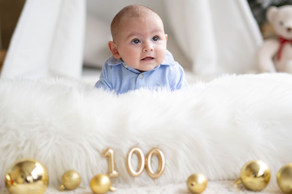 Baby in light blue outfit celebrates 100 day milestone with gold number balloons and decorations on white fur backdrop.