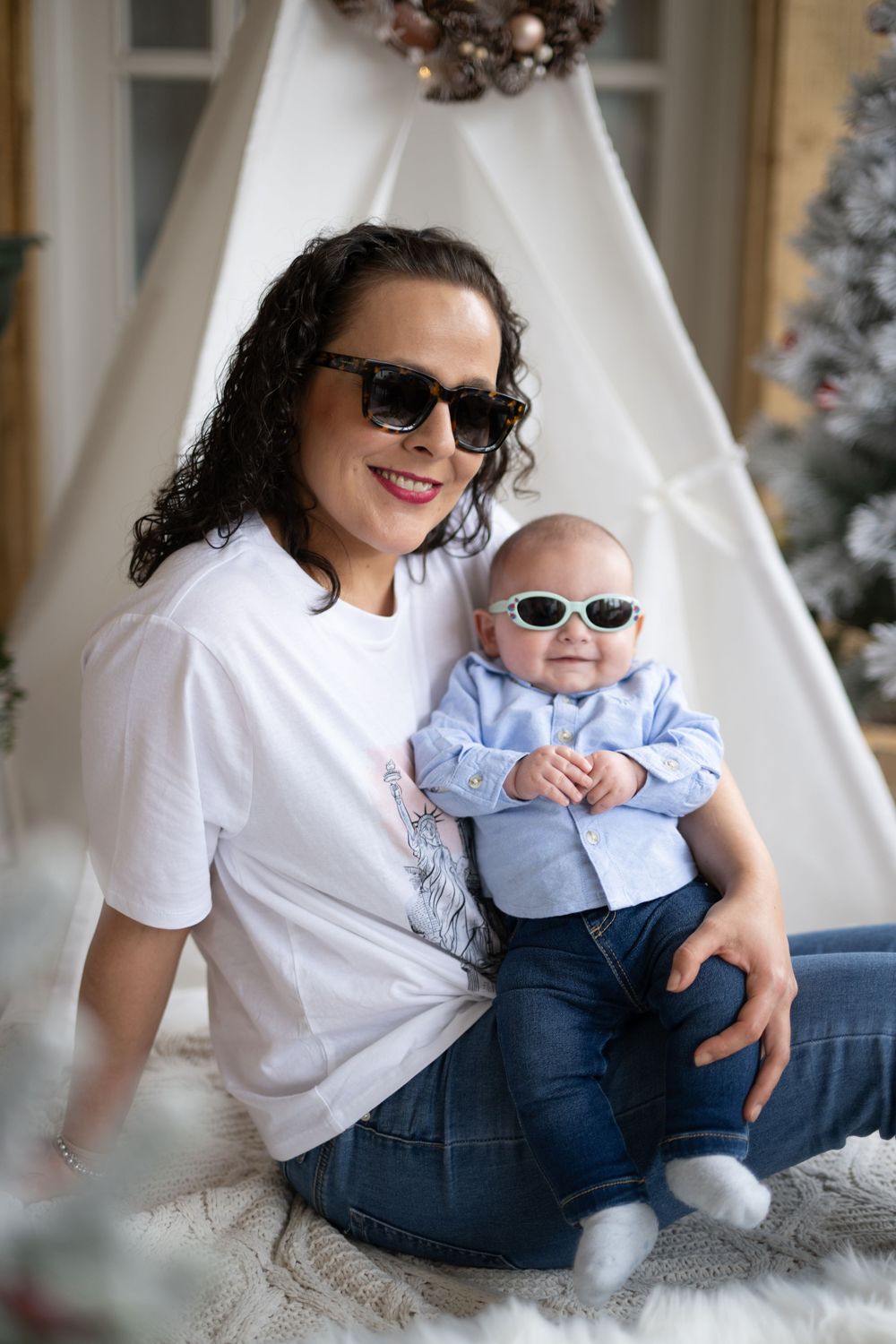 Parent and baby in matching white tops and sunglasses sit together on floor by Christmas decorations.