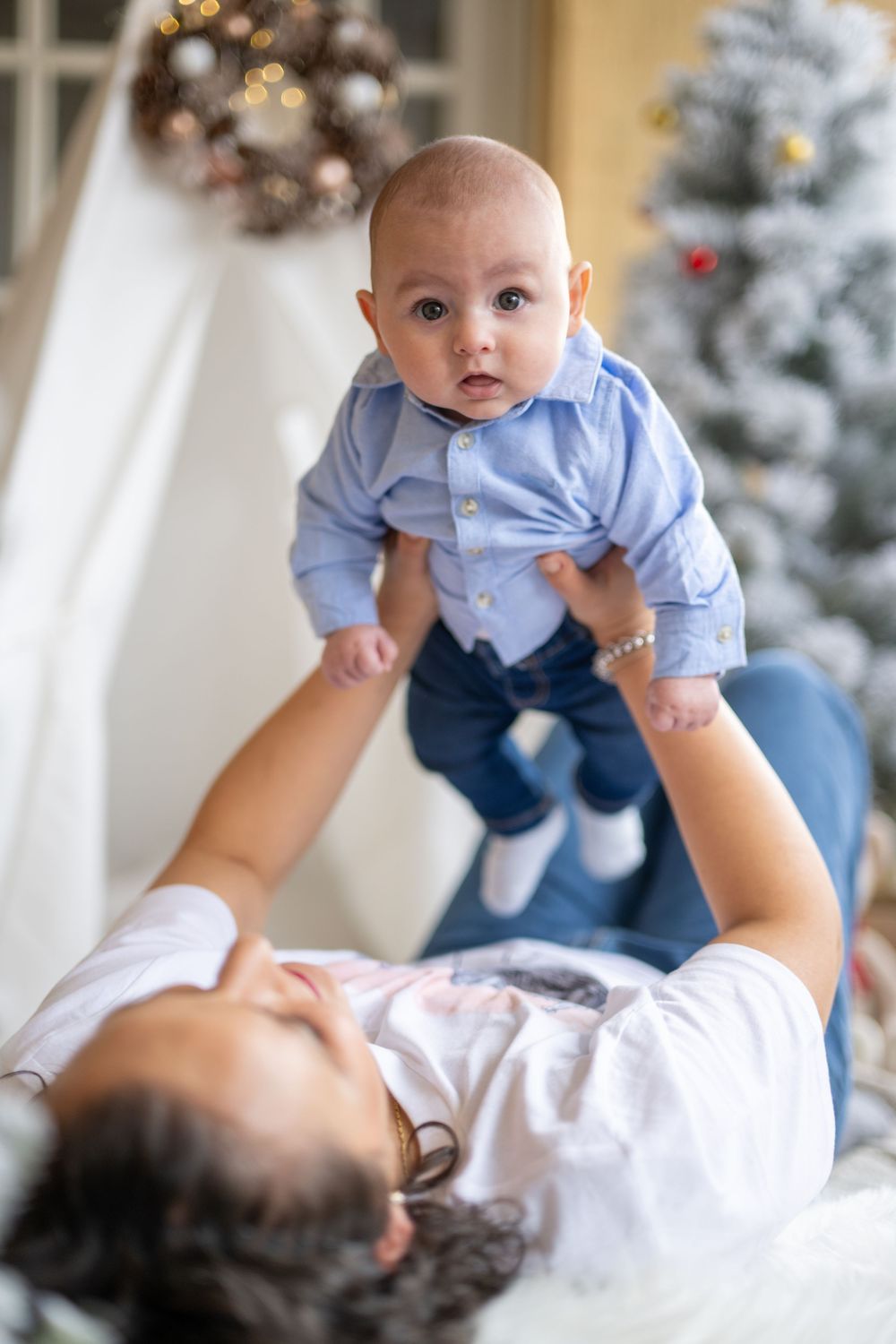 Baby in light blue shirt and jeans being lifted up in playful moment with holiday decorations in background.