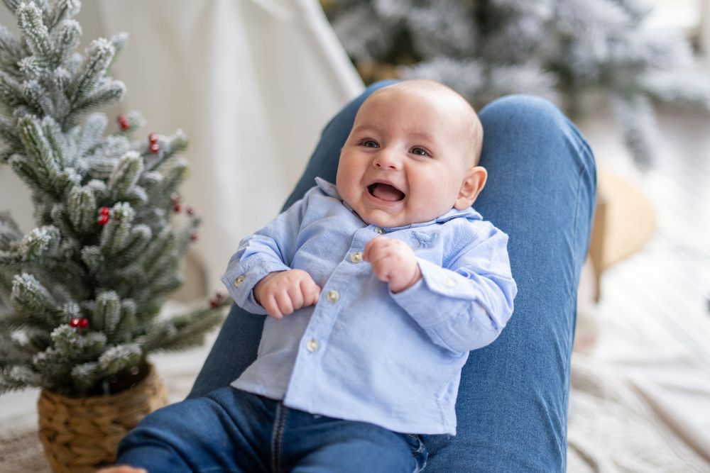 Baby in light blue outfit sits on couch during winter holiday photoshoot with decorated Christmas tree in background.