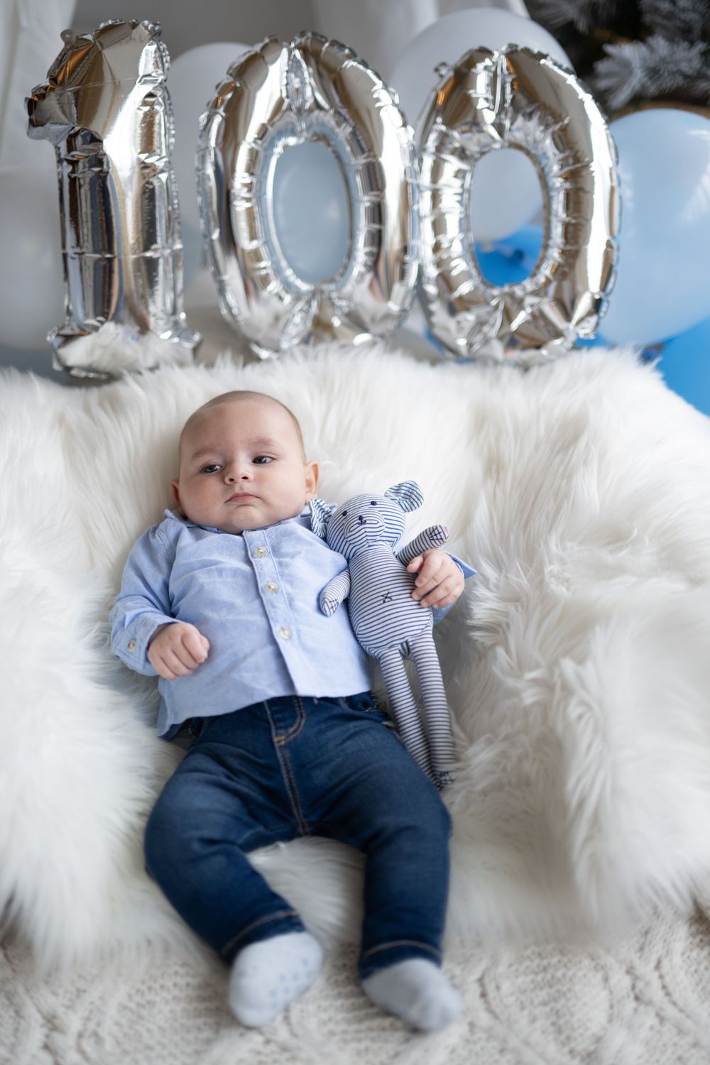 Baby lying on white furry blanket with silver number balloons in background.