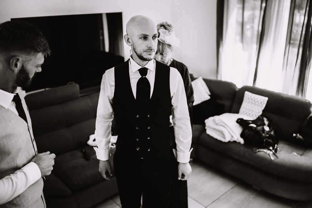 A black and white photo shows a man in a formal vest and tie standing in a room with leather couches.
