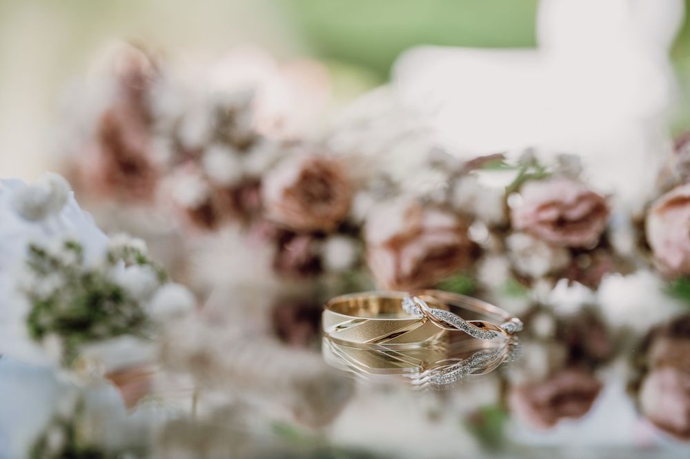 Wedding rings displayed on a soft surface with blurred pink roses and delicate floral arrangements in the background.