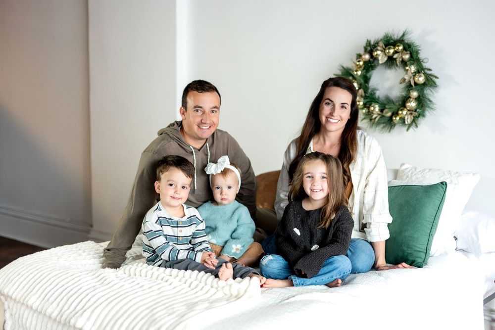 Family of five sitting on a bed with a gold Christmas wreath and festive holiday decor.