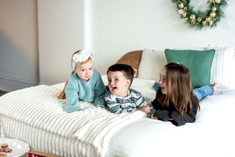 Three young siblings laugh and play together on a cozy white bed with a decorative Christmas wreath hanging on the wall.