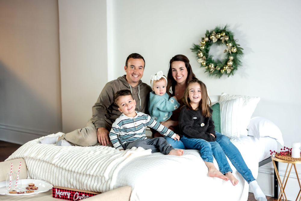 A family of five sits together on a bed, smiling for a festive Christmas portrait with a holiday wreath on the wall.