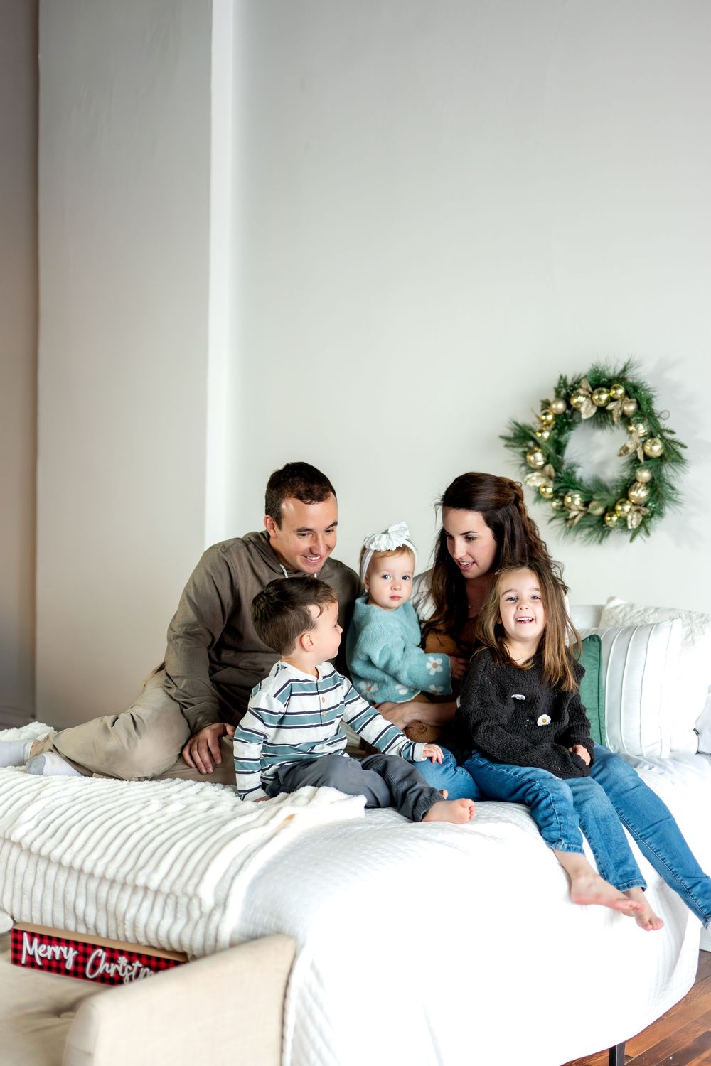 A happy family of five sits on a white bed, sharing a joyful moment together with a Christmas wreath on the wall.