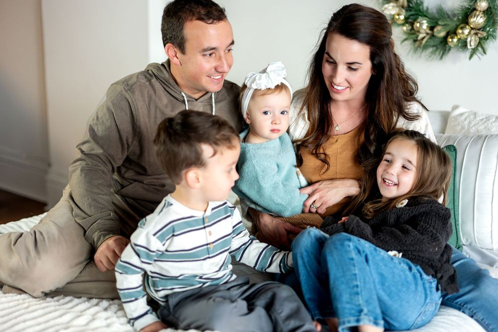 A loving family of five sits closely together on a bed, laughing and interacting with a Christmas wreath in the background.