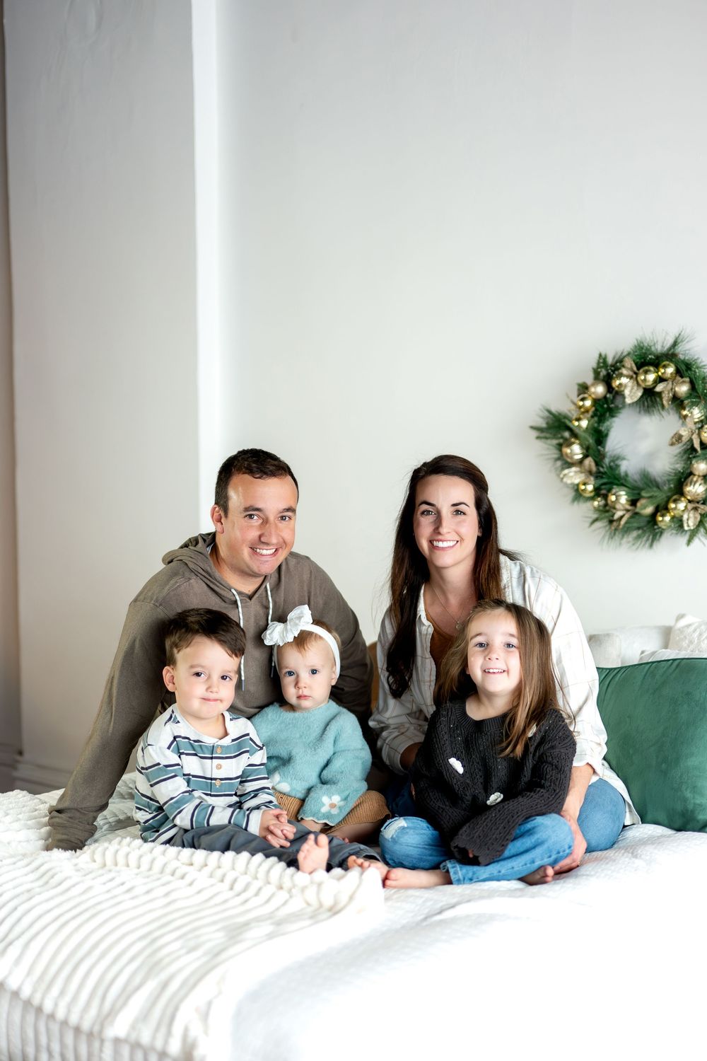 Smiling family of five posing on a white bed with a decorative Christmas wreath on the wall.