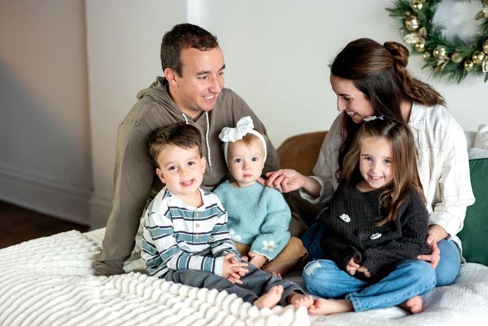 Family of five laughing together on a white bed with a gold holiday wreath in the background.