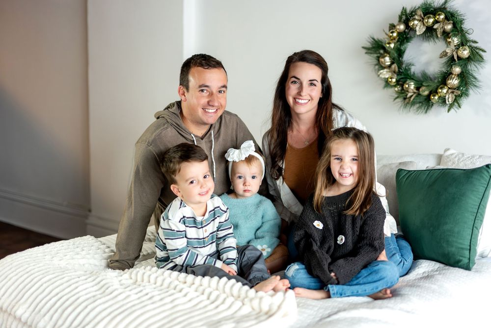 Family of five posing on a white bed with a festive gold Christmas wreath on the wall.