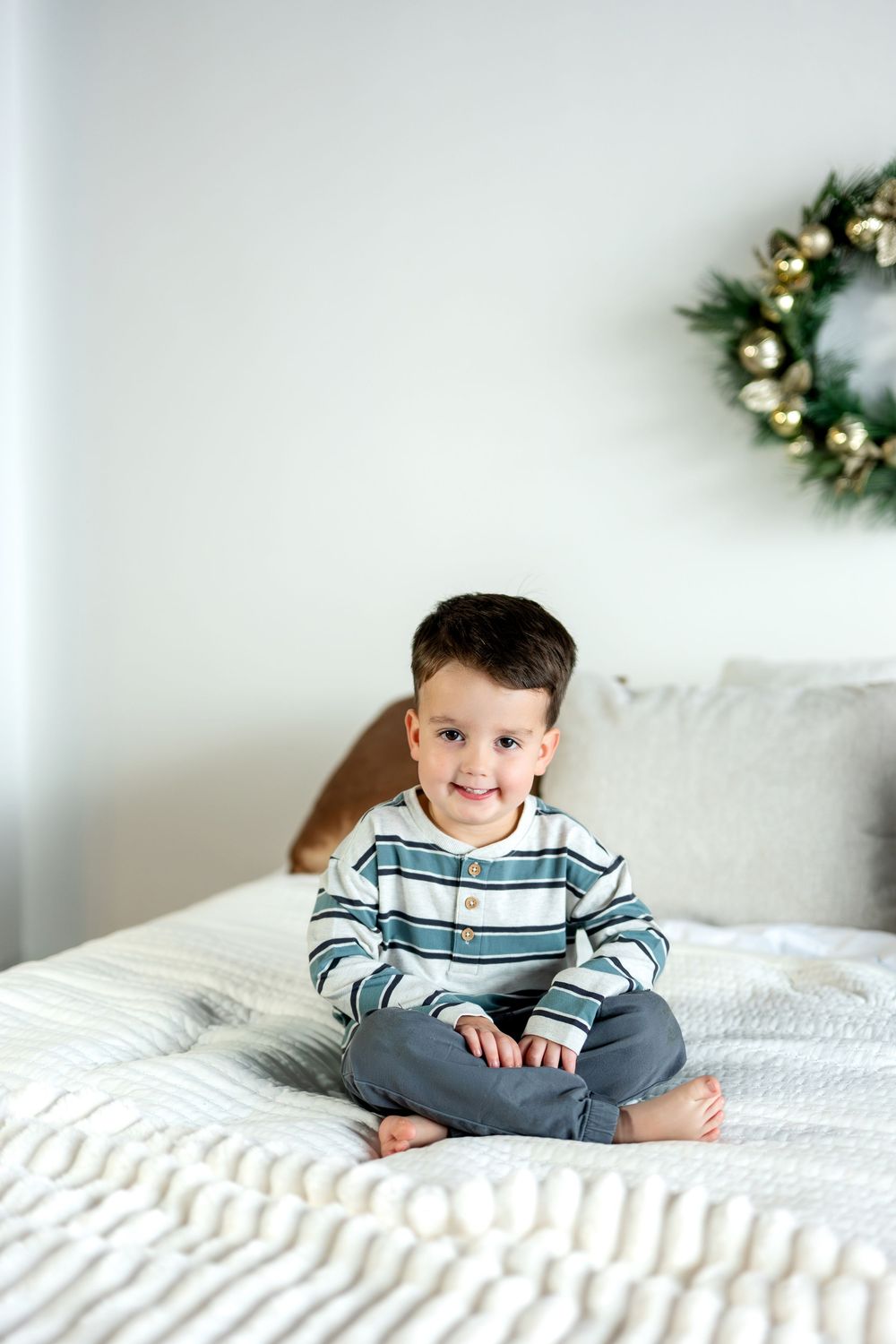 Adorable young boy sitting cross-legged on a white bed with a Christmas wreath behind him.
