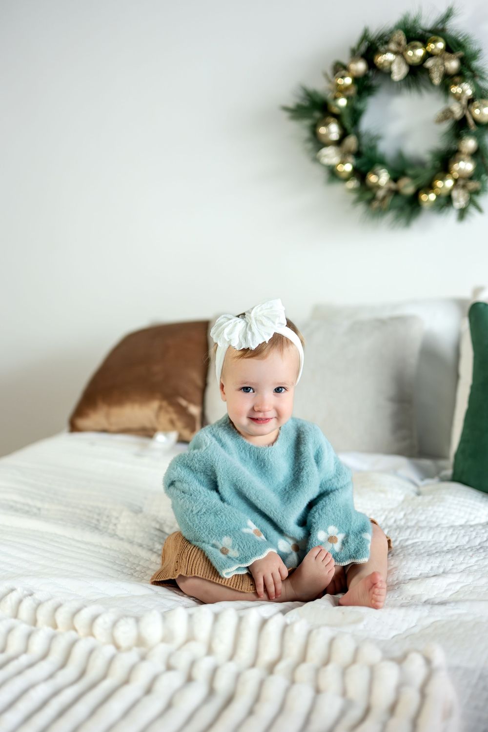 Cute baby girl in a teal sweater and white bow headband sitting on a bed with holiday decor.