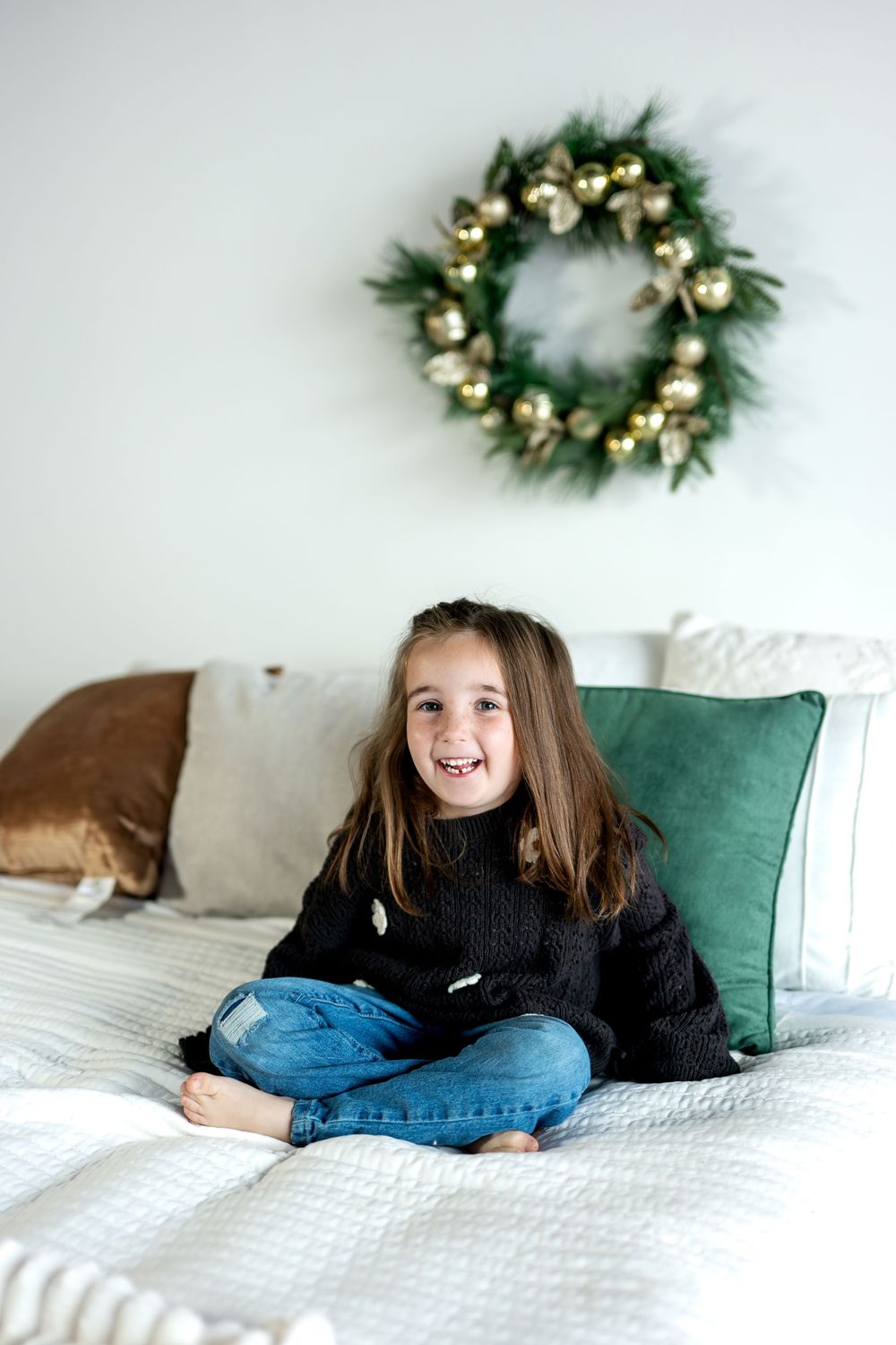 A happy young girl sits cross-legged on a bed in a black sweater and jeans, smiling with a Christmas wreath behind her.