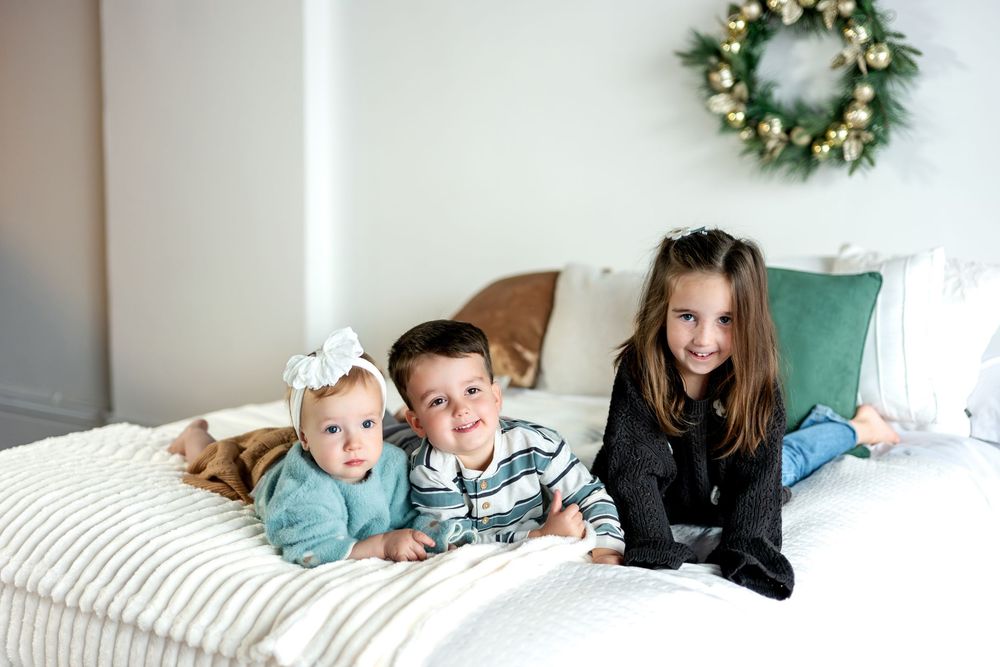 Three young siblings lie together on a white bed, smiling, with a festive Christmas wreath decorating the wall behind them.