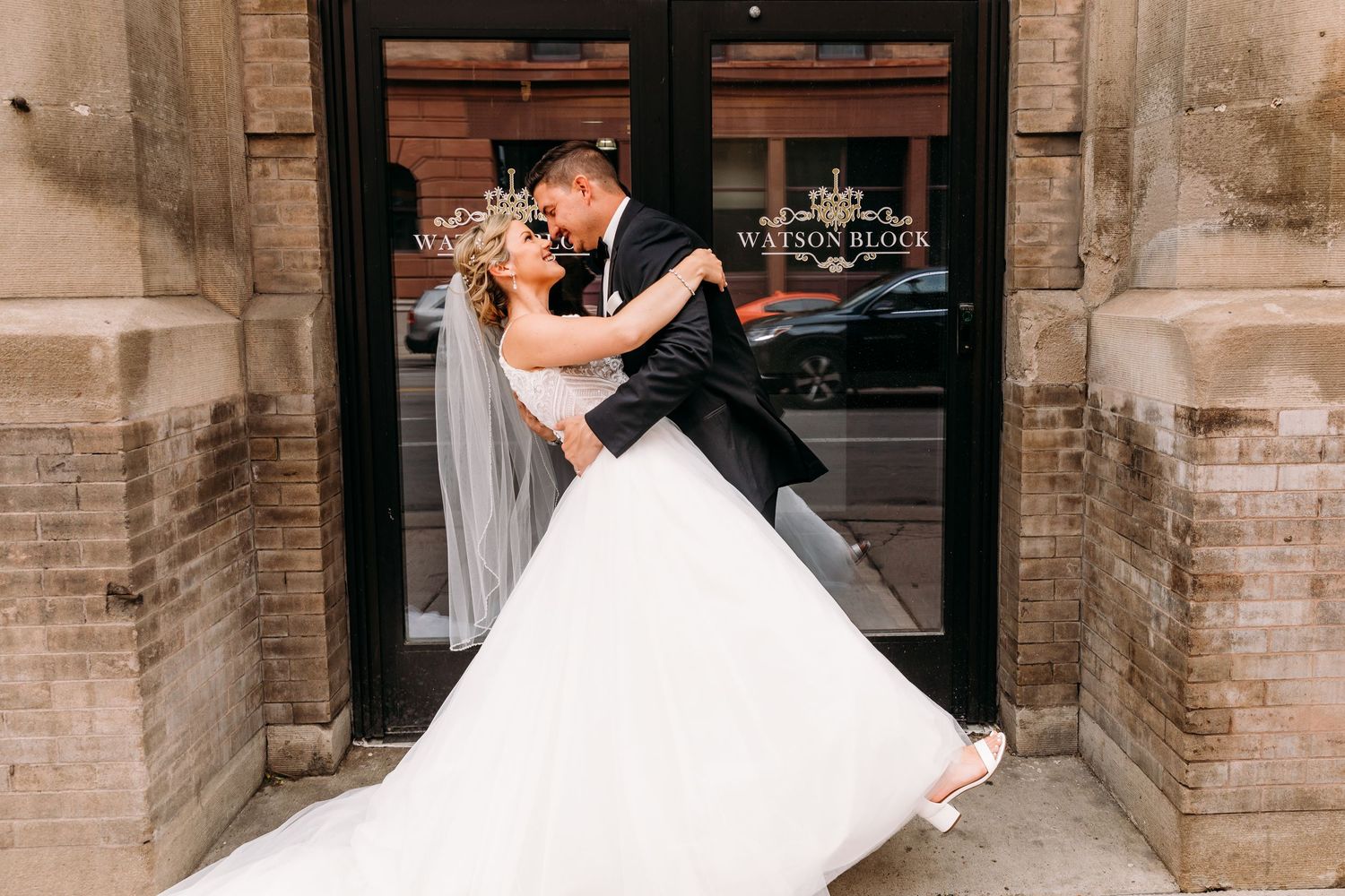 A romantic wedding couple dances and embraces in front of a rustic brick building entrance.