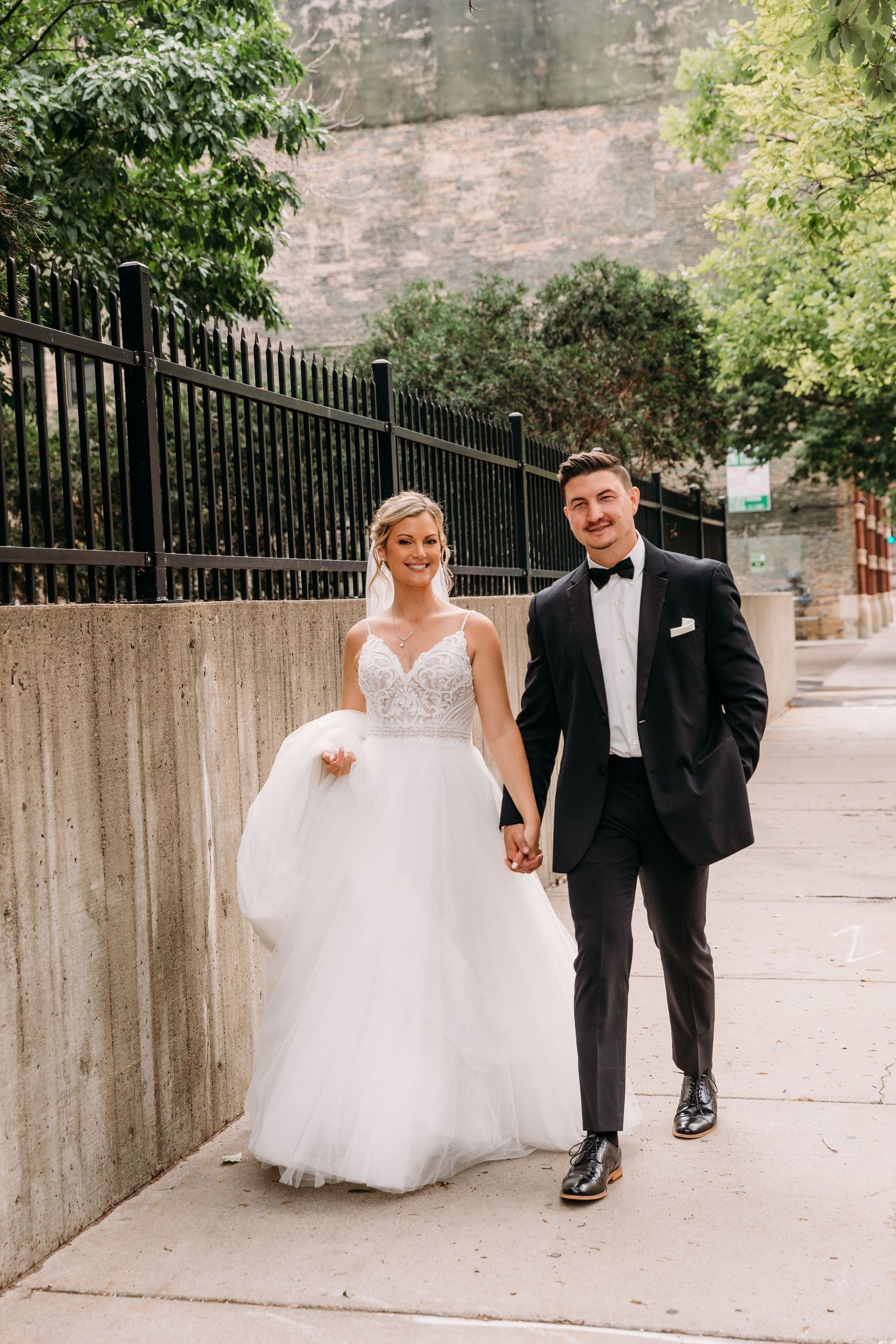 A sequence of wedding photos showing a couple walking along a sidewalk with a brick wall and iron fence.