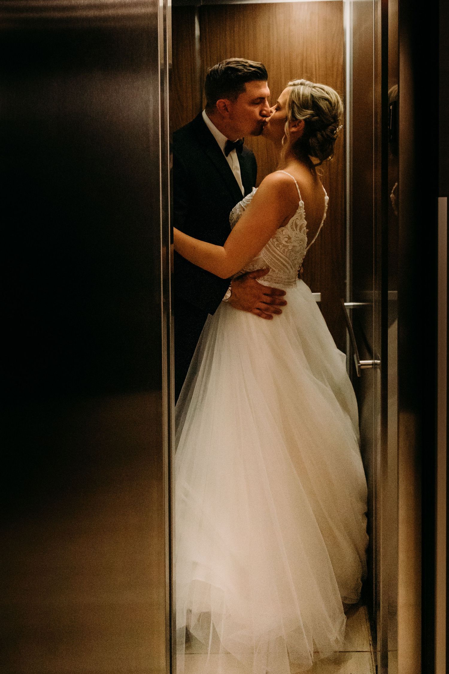A romantic wedding couple shares an intimate moment in a dimly lit elevator, with the bride wearing a flowing white gown.