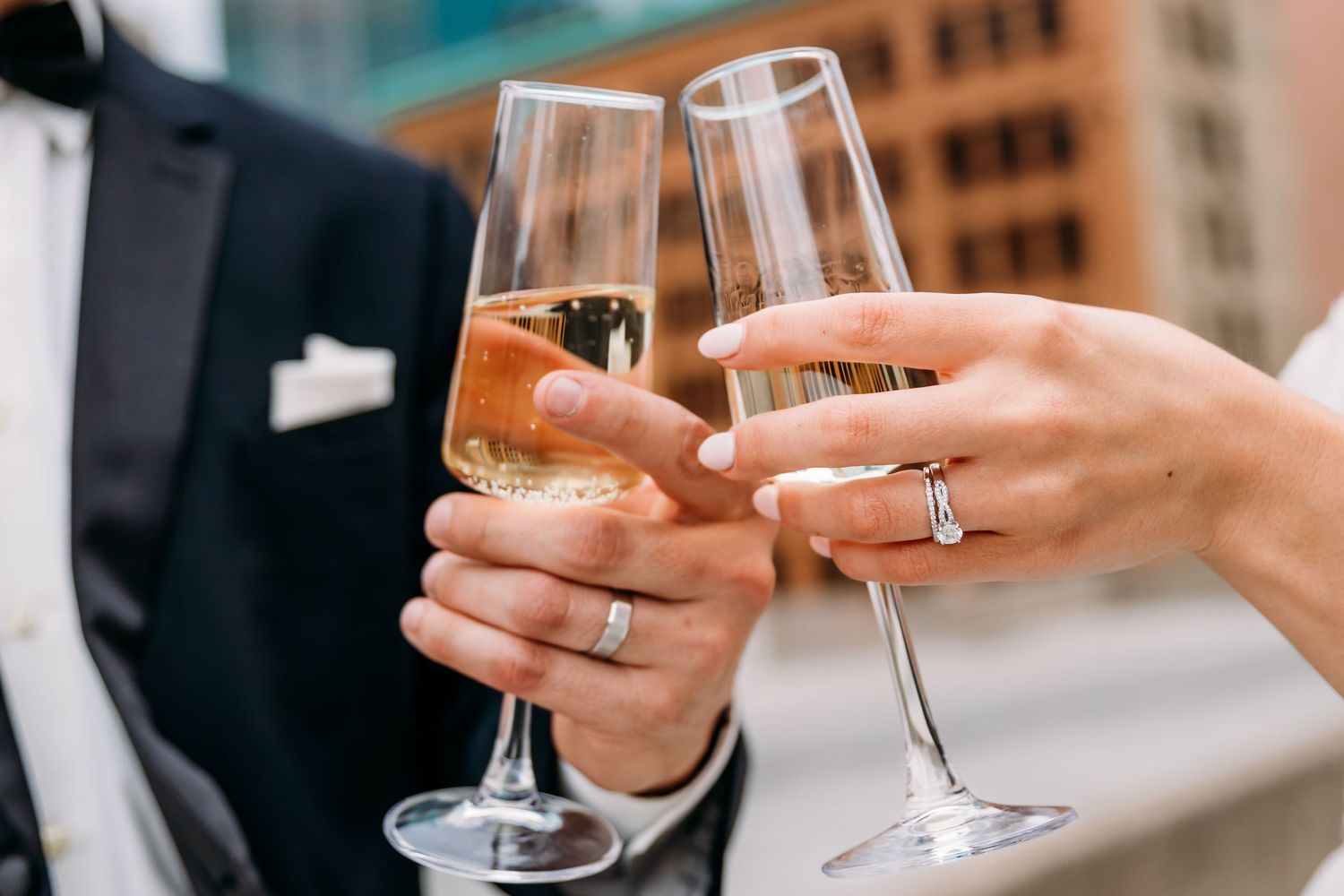 Two hands clink champagne glasses in a celebratory toast with city buildings in the background.