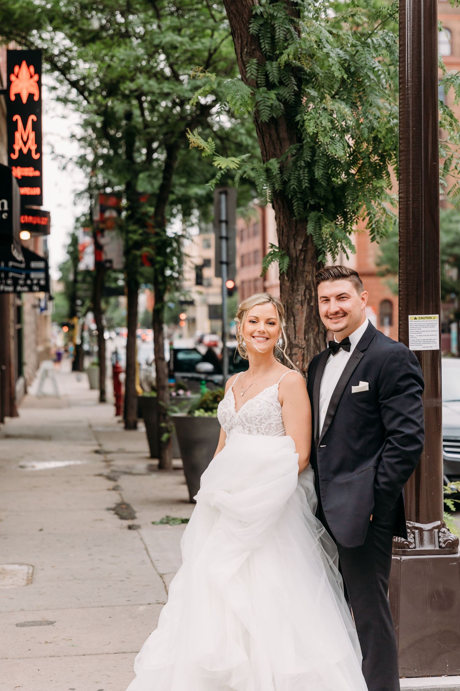 A wedding couple poses on a city sidewalk with trees and a neon sign visible in the urban background.