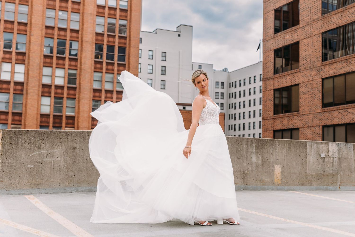A flowing white wedding dress billows dramatically against an urban rooftop backdrop with brick buildings in New York City.