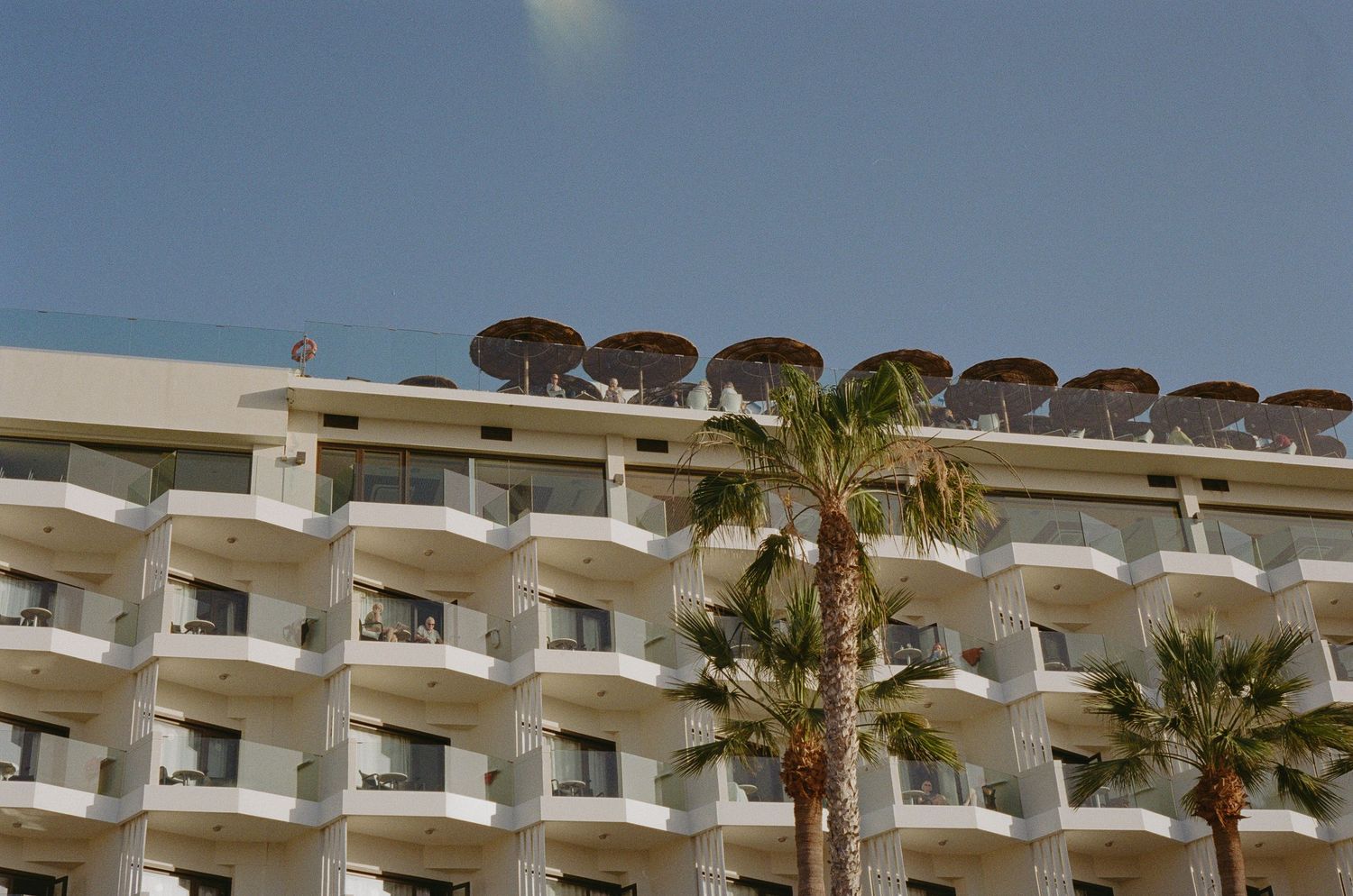 Pink apartment buildings perched on rocky cliffs along a Mediterranean coastline under bright blue skies.