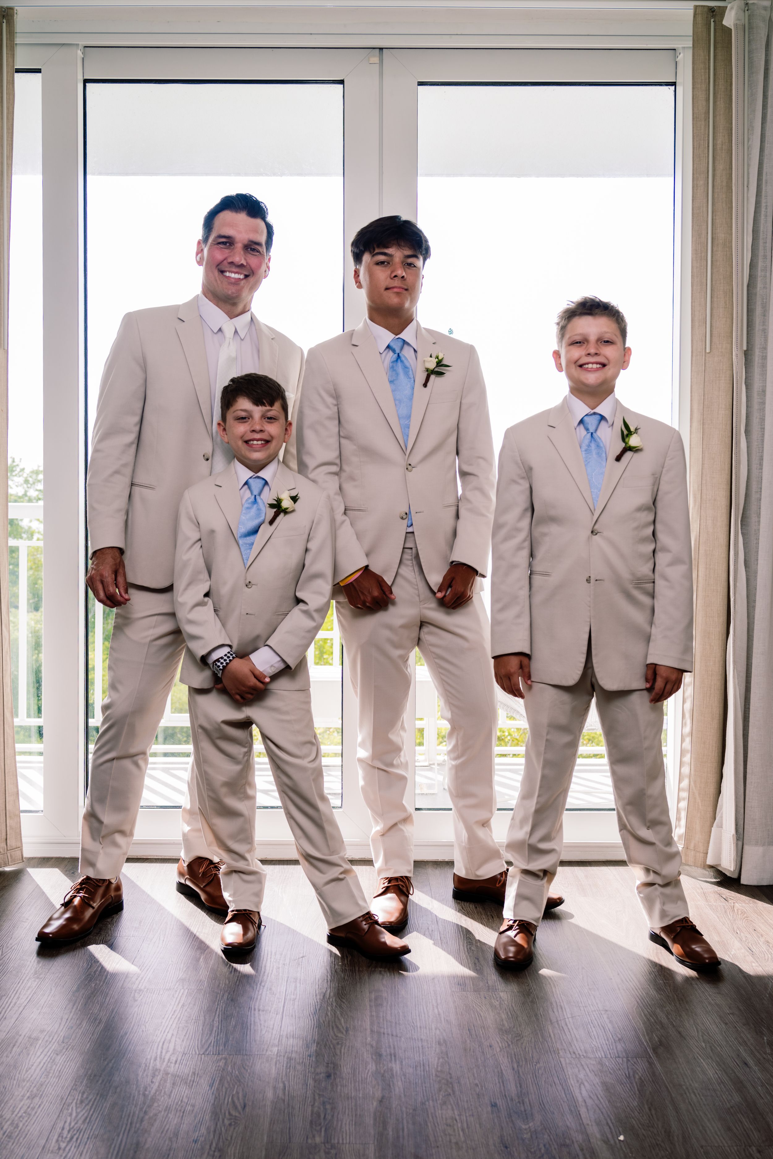Four males in matching tan suits with blue ties and brown loafers pose together at a wedding celebration. Groom Kevin with his three sons in