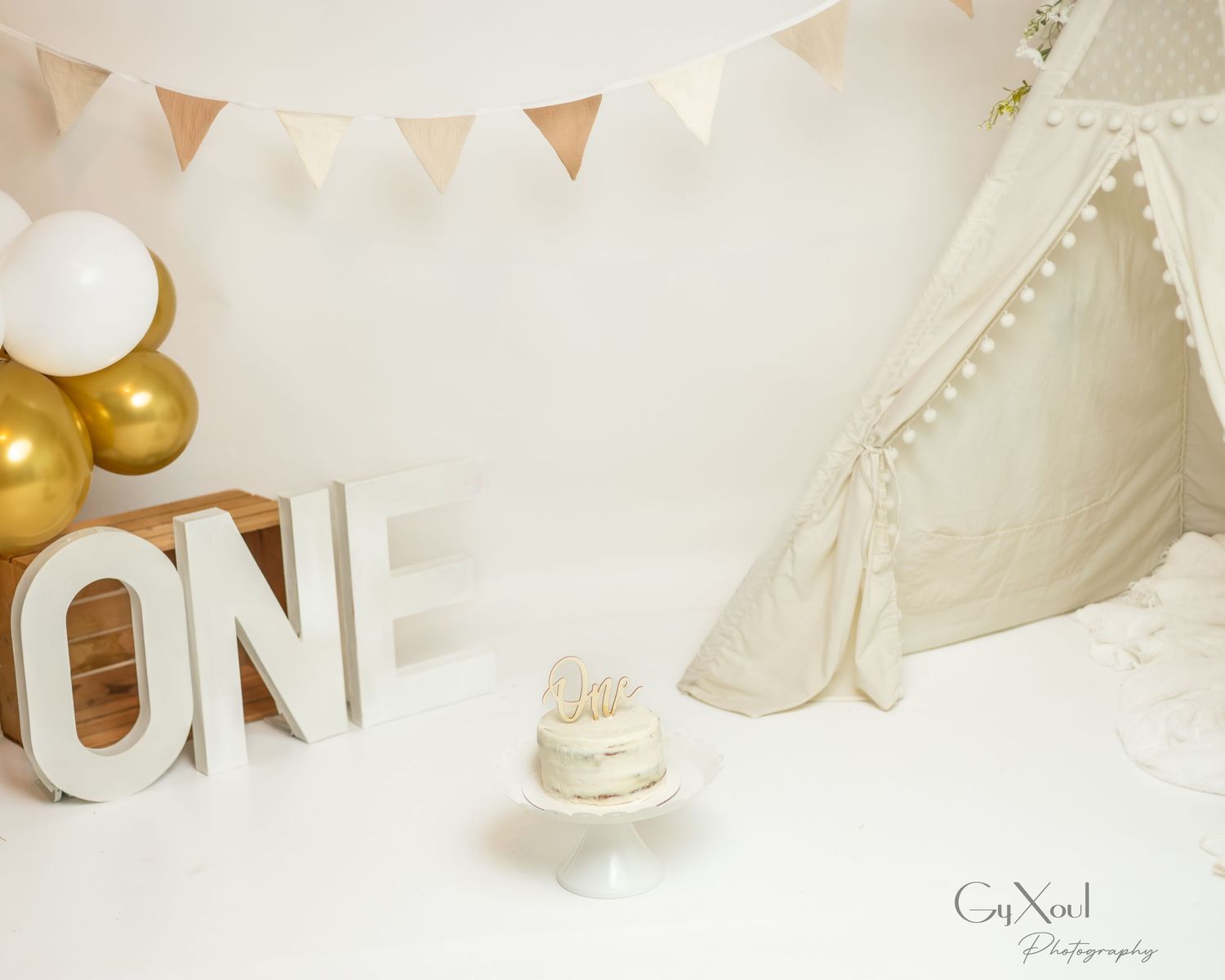 First birthday cake smash setup with ONE letters, white teepee, gold and white balloons, bunting, and small cake on white backdrop.