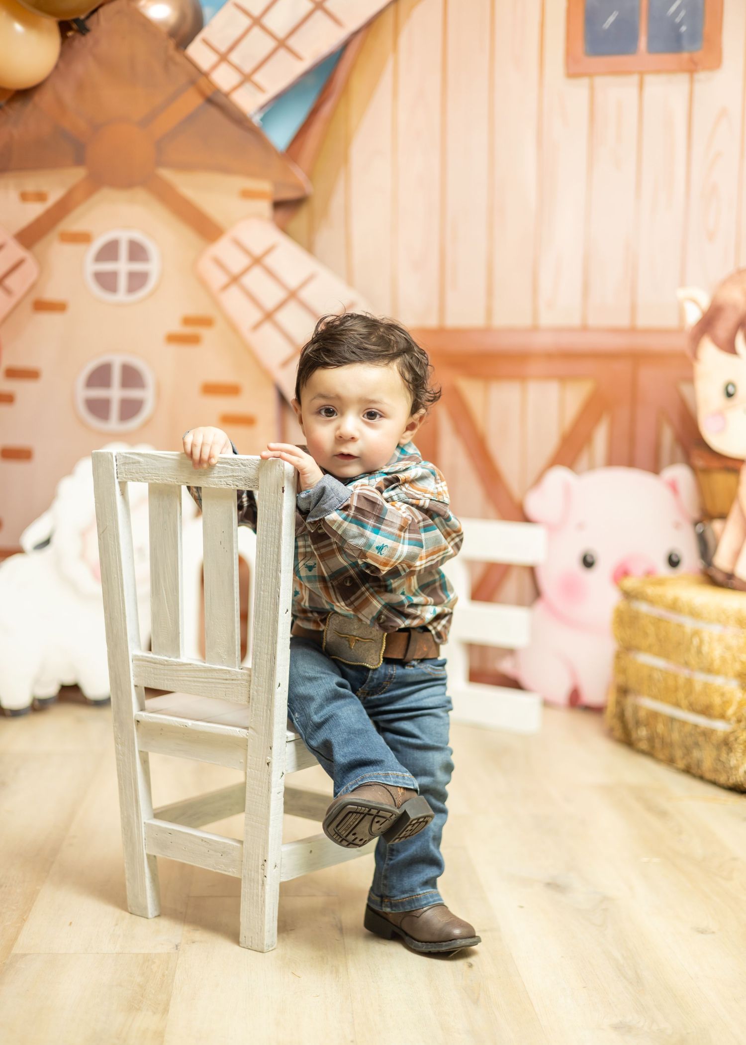 Toddler boy in plaid shirt and jeans leaning on white chair at farm-themed birthday party backdrop.