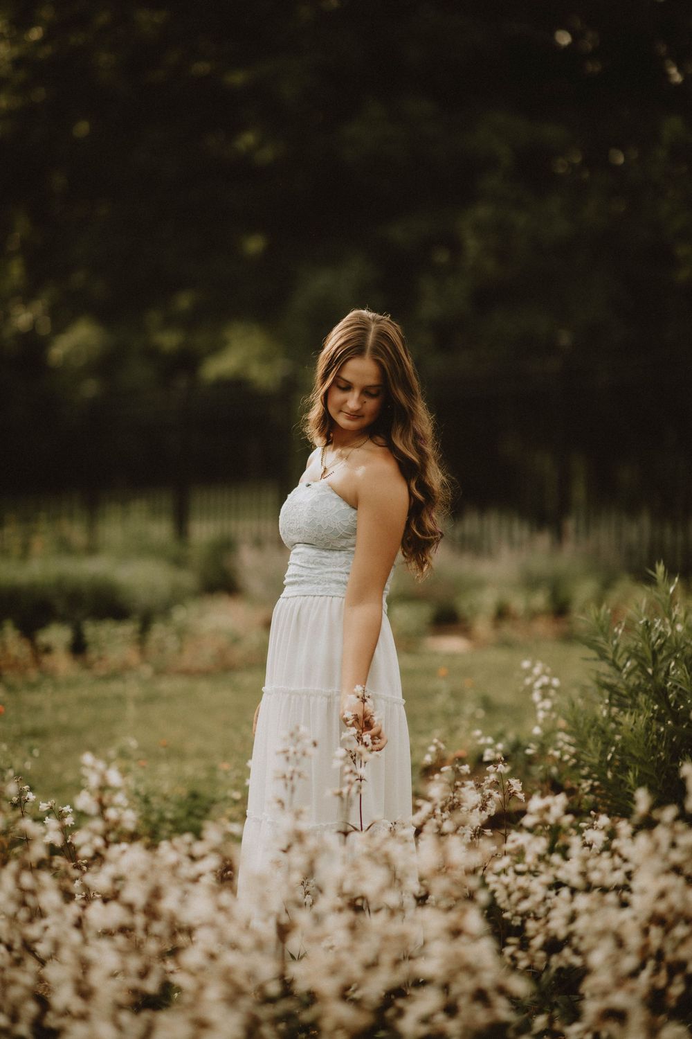 Young woman in white dress standing among wildflowers in a garden, looking down with long wavy hair. Pittsburgh PA Senior Photographer