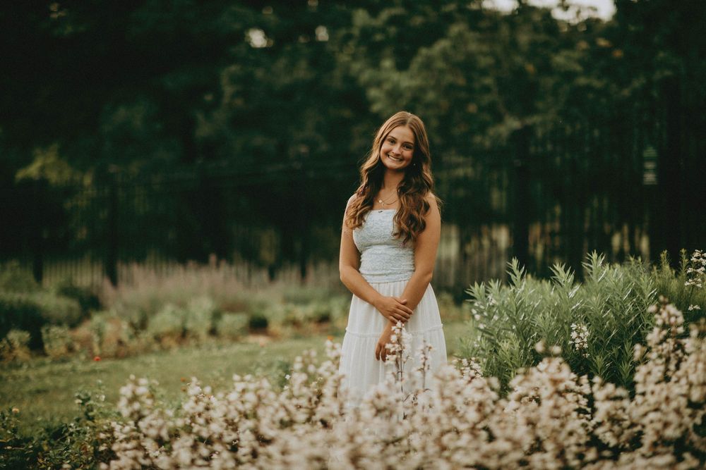 Young woman in white dress smiling among wildflowers in a lush garden setting. hartwood acers senior portraits pittsburgh photographer